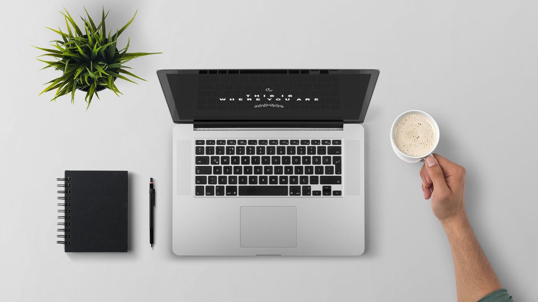 A laptop on a desk beside a plant, a notebook and a cup of coffee